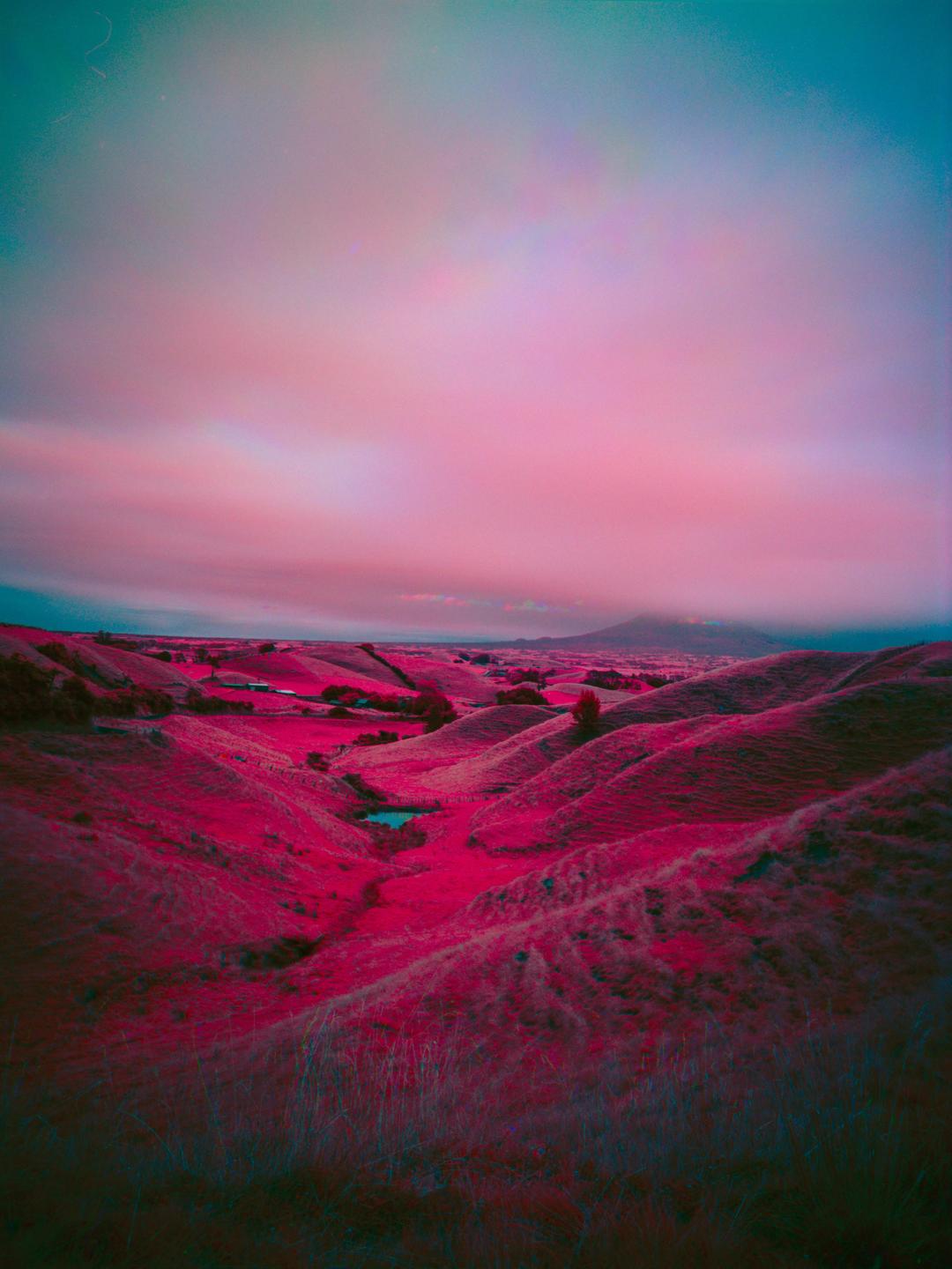 A landscape of rolling hills with a small pond in a valley is visible under a pink and blue sky.