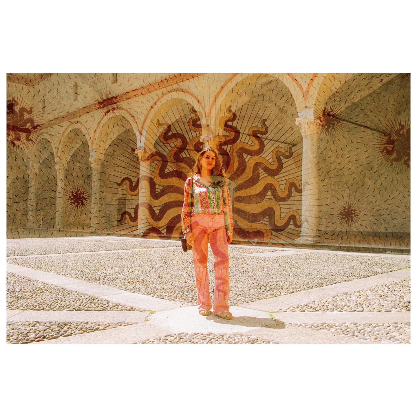 A woman stands in a courtyard with arched walls and a patterned floor, with a sun-like design superimposed on the background.