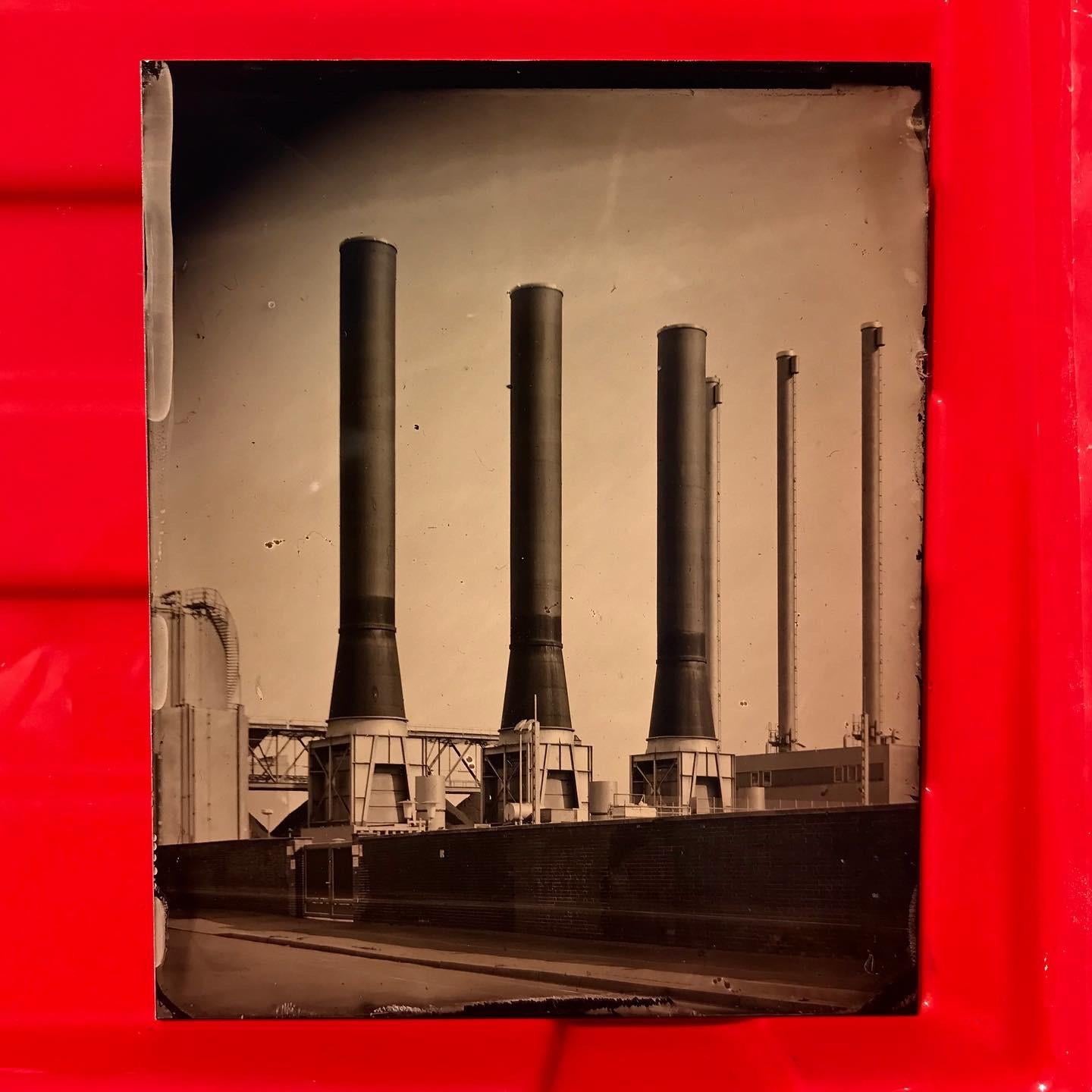 Four tall industrial smokestacks rise above a building with a road in front of it.