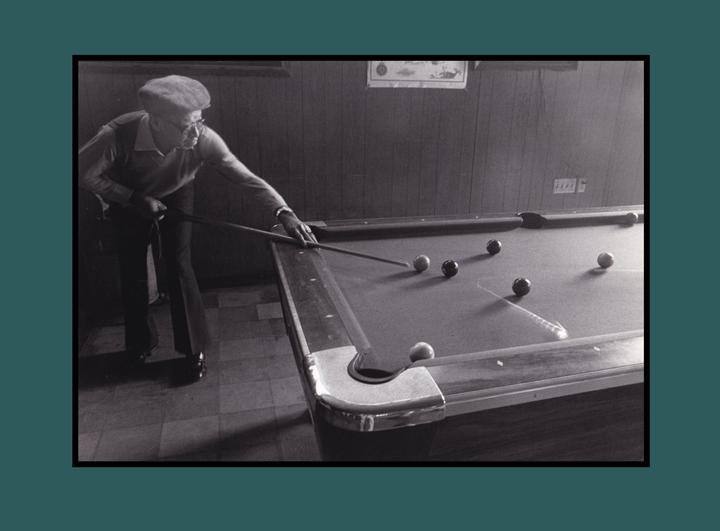 A man in a flat cap leans over a pool table, cue stick in hand, aiming at a cue ball.