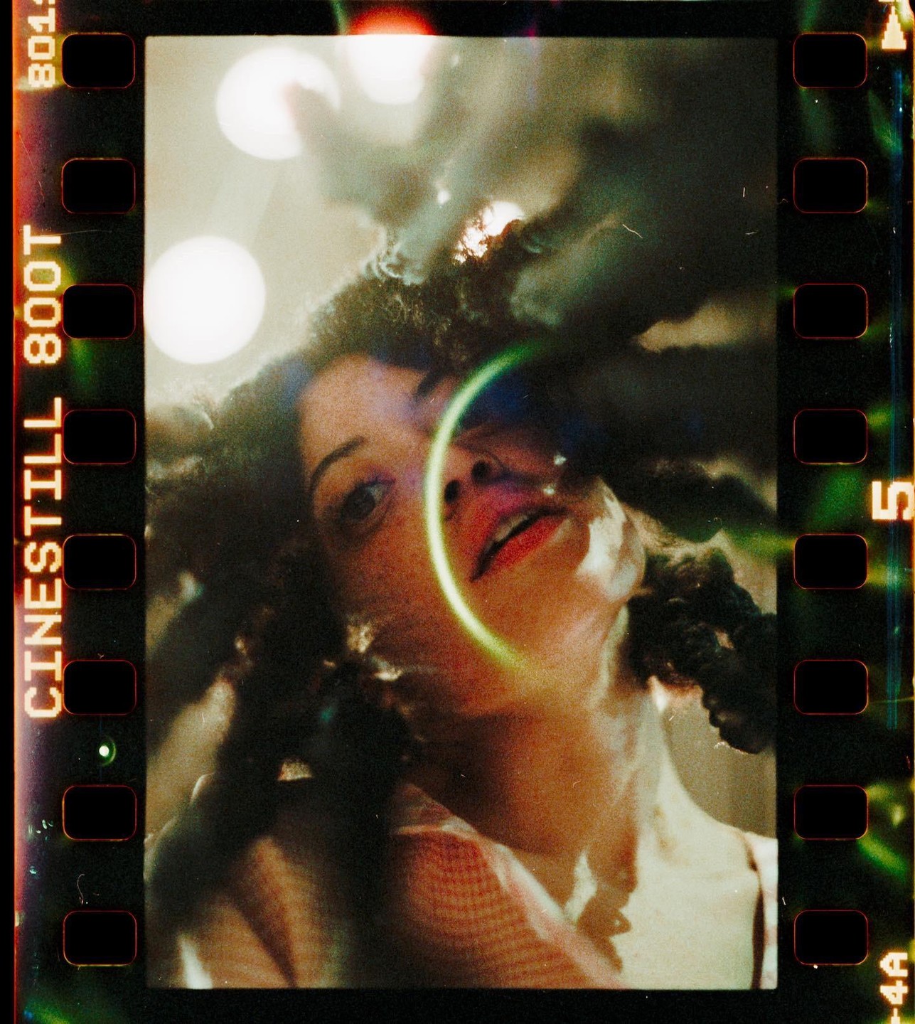 A woman with curly hair looks up, with a bright green light ring around her face and bokeh lights in the background.