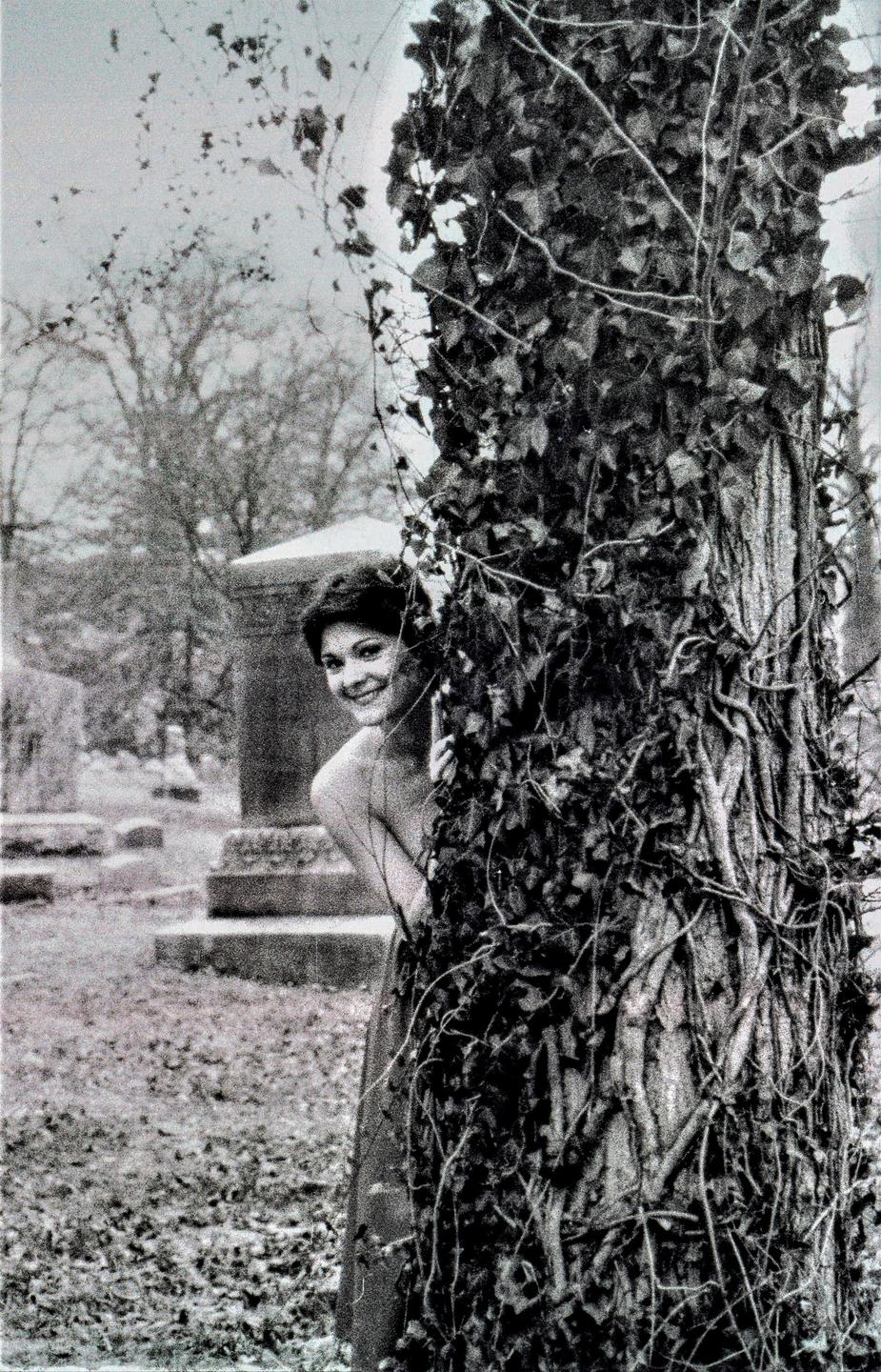 A woman peeks from behind an ivy-covered tree in a cemetery.