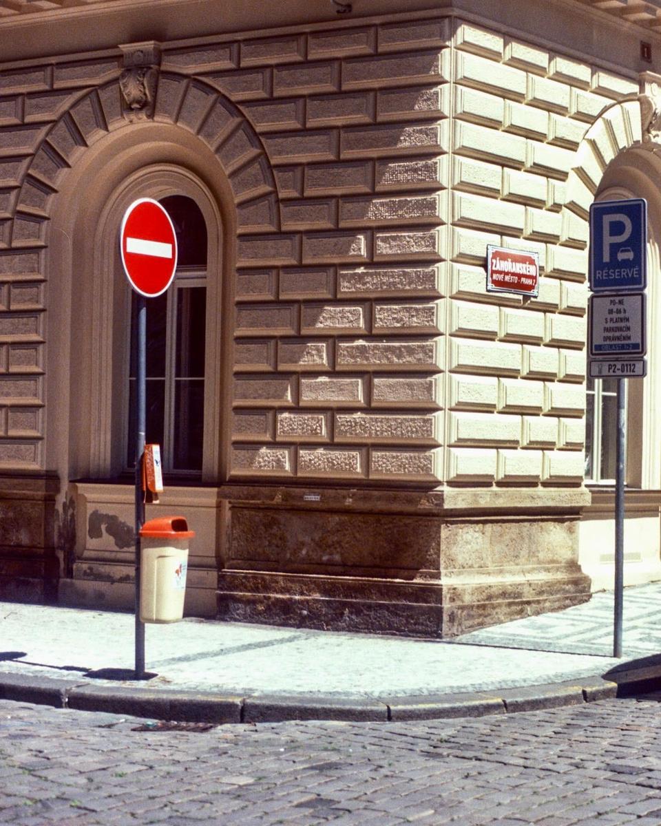 A no entry sign is attached to a pole next to a building with textured stone walls and arched windows, with a parking sign and a trash can also visible.