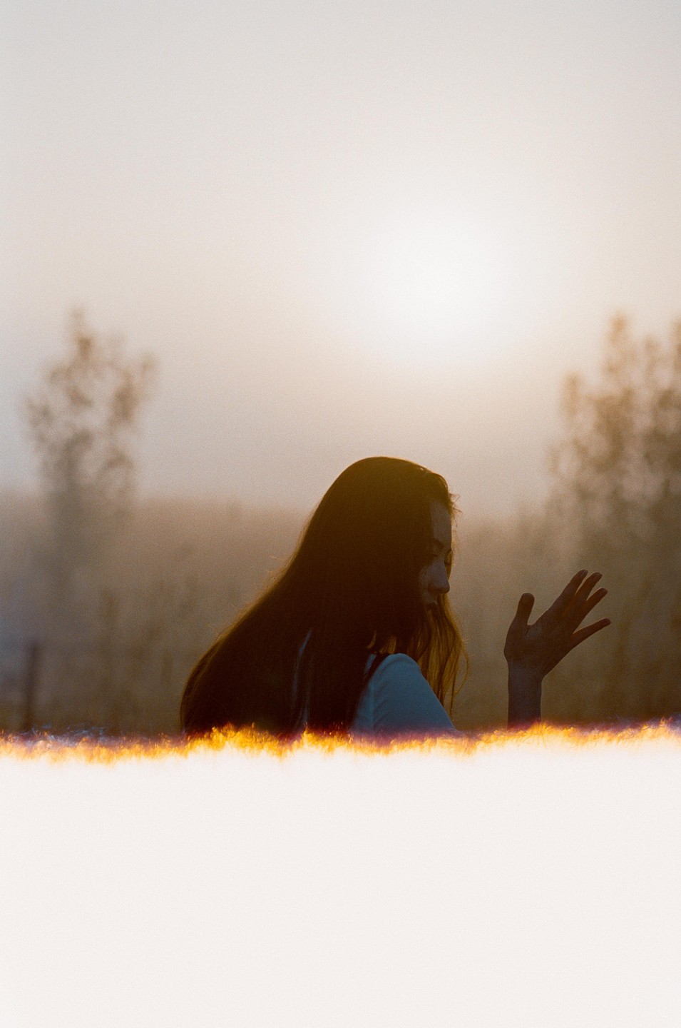 A woman with long hair is silhouetted against a hazy, bright sky with a glowing light leak at the bottom.