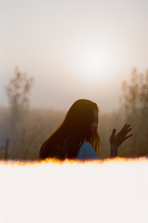 A woman with long hair is silhouetted against a hazy, bright sky with a glowing light leak at the bottom.