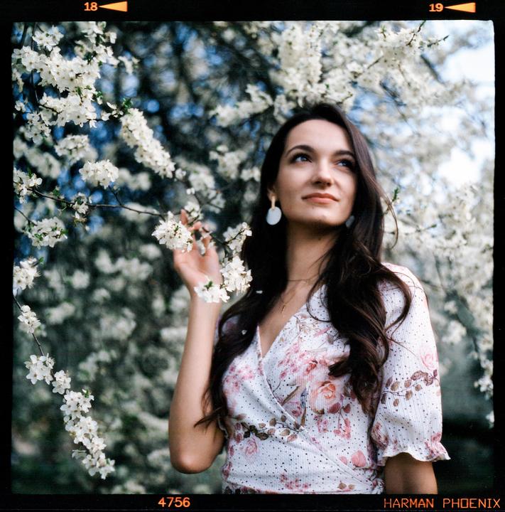 A woman with long dark hair and a floral dress stands among white blossoms, looking upwards.
