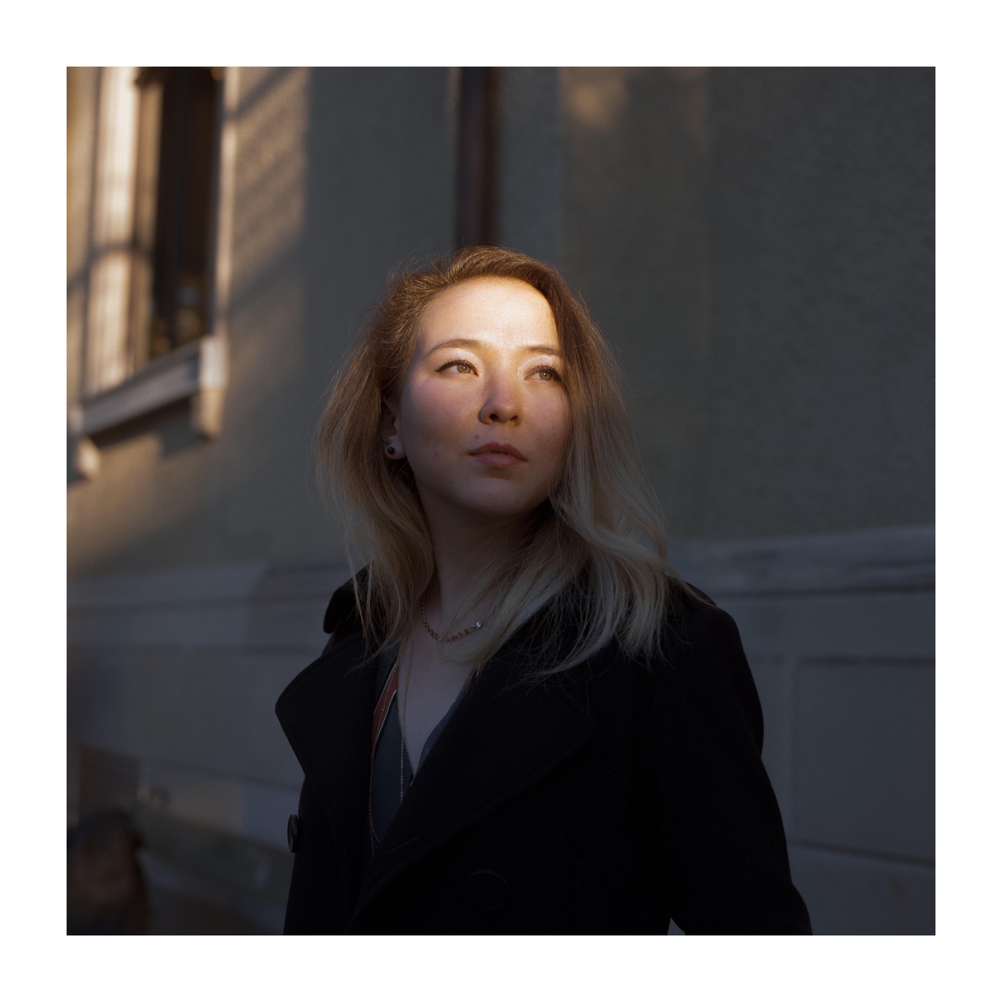 A woman with blonde hair looks up, illuminated by a patch of sunlight on her face, with a building in the background.