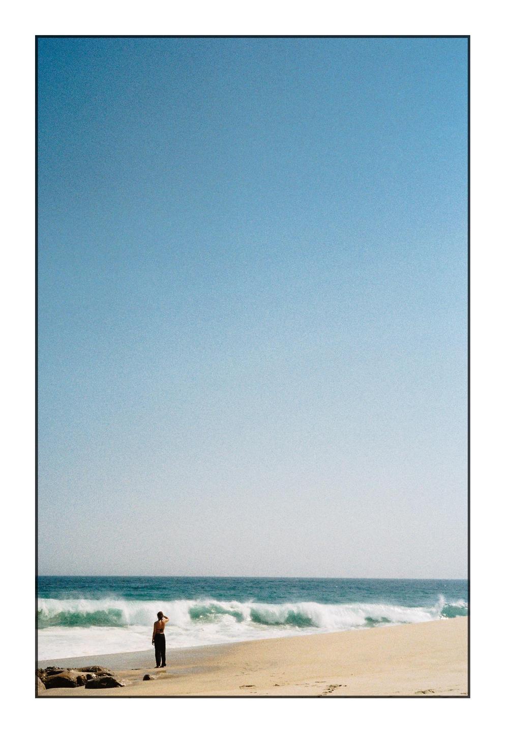 A person stands on a sandy beach looking out at the ocean waves under a clear sky.