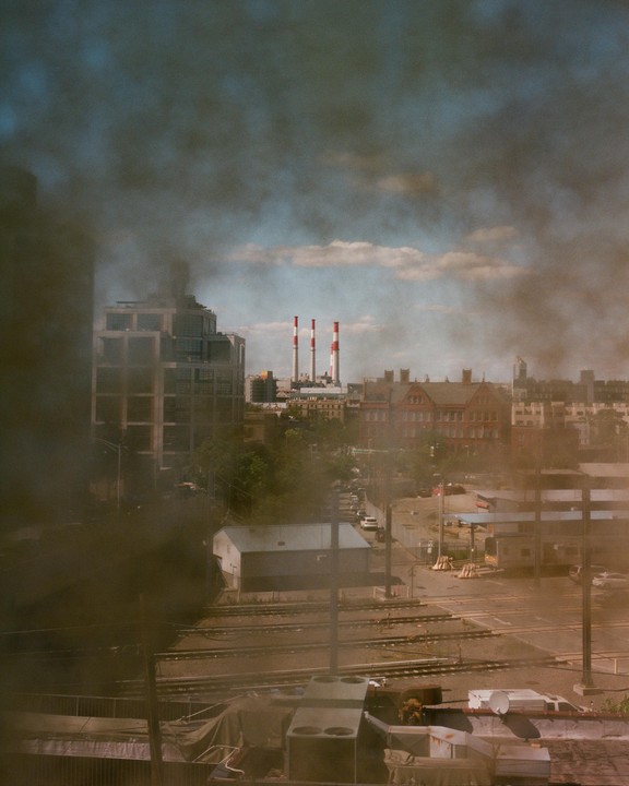 Smoke billows over a city with industrial smokestacks in the distance.