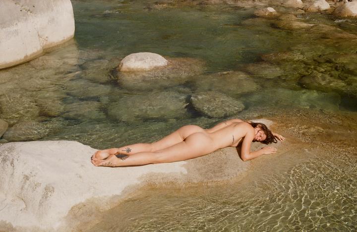 A woman lies nude on a rock next to clear water.