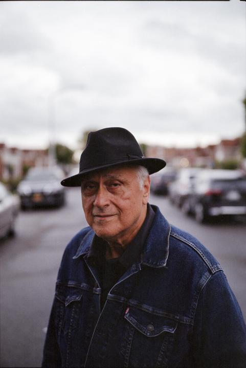 A man wearing a hat and a denim jacket stands on a street with cars parked on both sides.