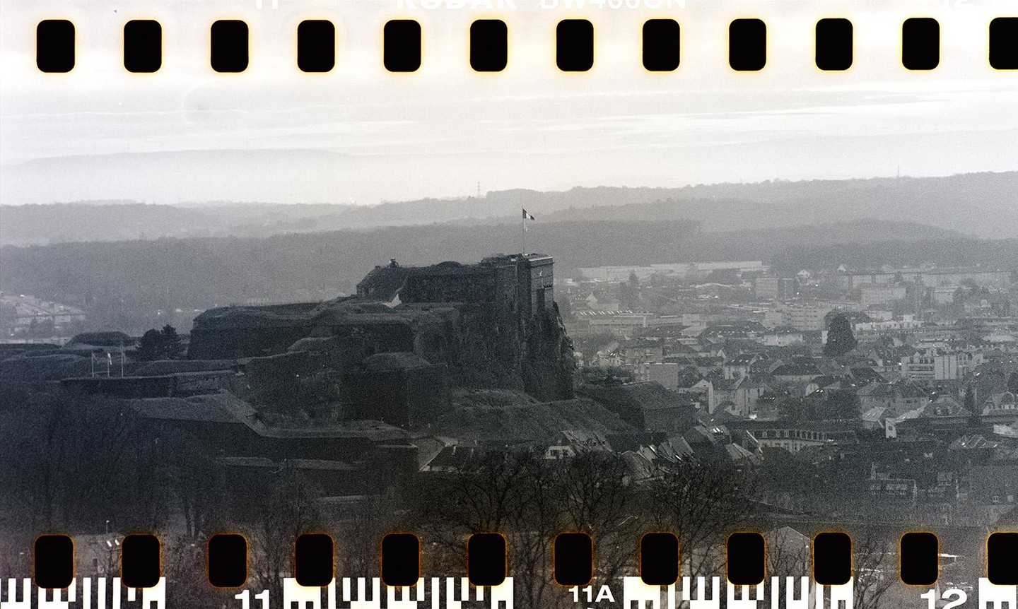 A citadel with a flag on top overlooks a town with many buildings and a hazy mountain range in the background.