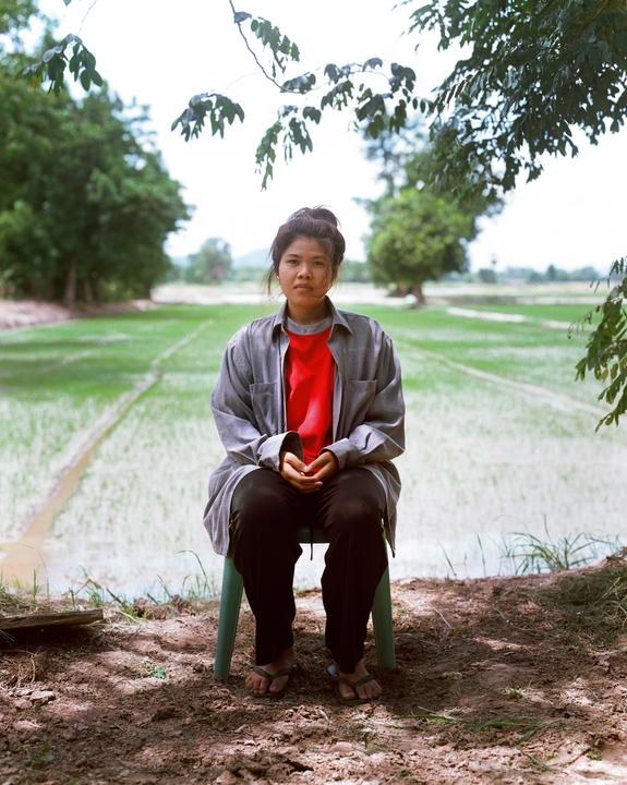 A woman sits on a chair under a tree with a rice paddy in the background.