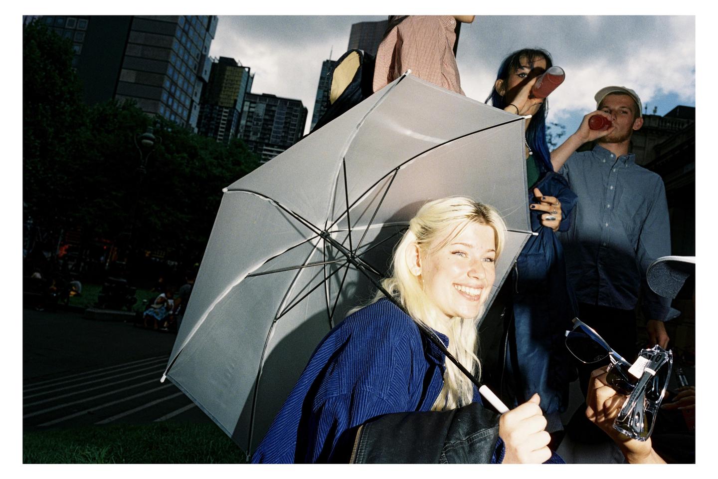 A woman smiles while holding an umbrella, with other people and buildings in the background.