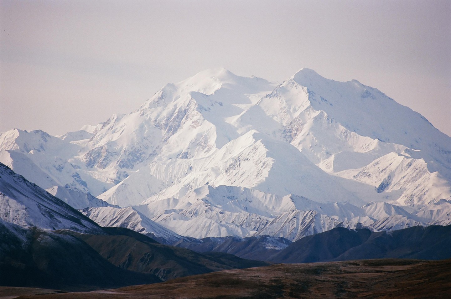 A vast snow-covered mountain range rises above a dark, barren landscape under a pale sky.