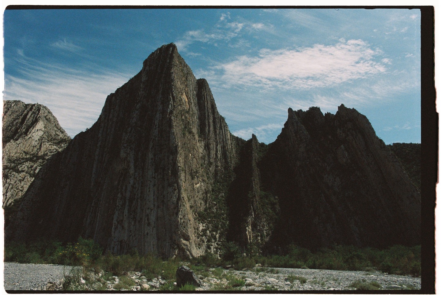 A wide shot shows a rocky riverbed in front of a large, jagged mountain range under a blue sky with wispy clouds.