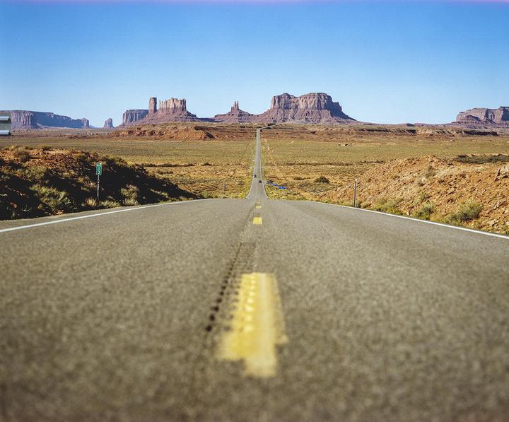 A long, straight road leads towards distant mesas and buttes under a clear sky.