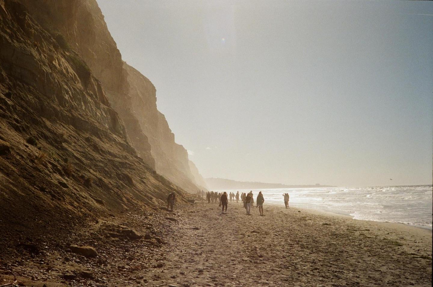 A group of people walk along a sandy beach next to towering cliffs and the ocean.