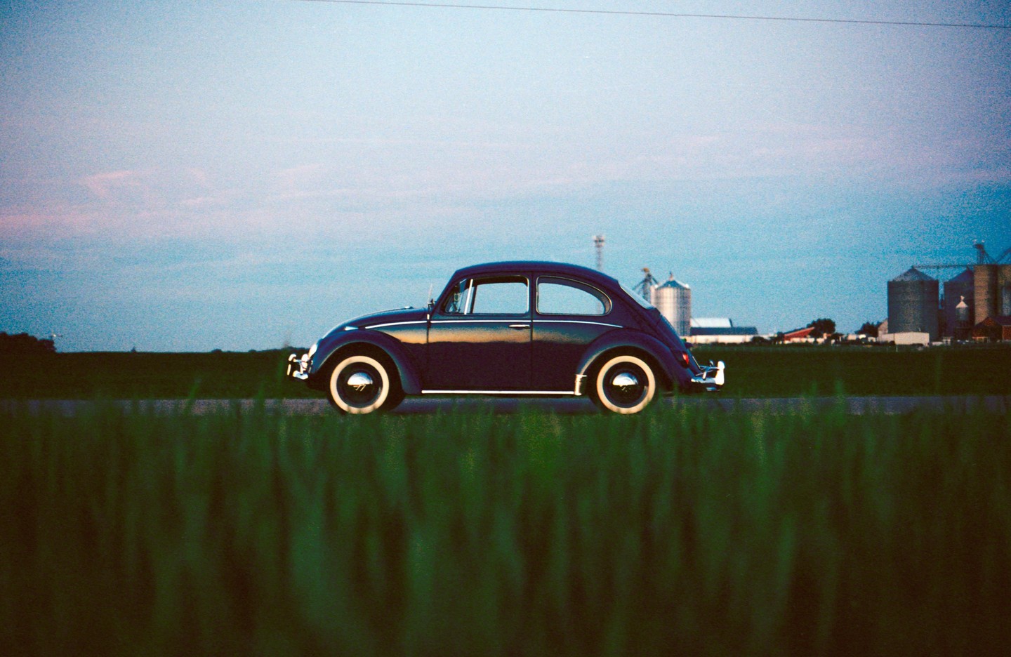 A dark blue vintage beetle car is parked on a road next to a field with grain silos in the distance under a blue sky.