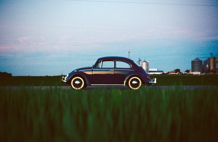 A dark blue vintage beetle car is parked on a road next to a field with grain silos in the distance under a blue sky.