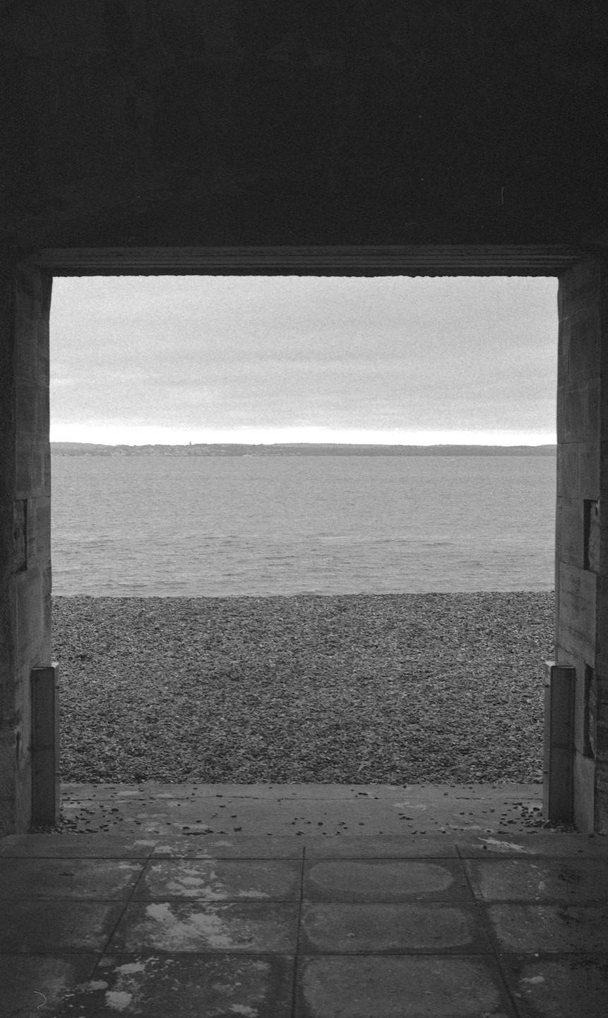 A view of the sea and a pebble beach is framed by a dark concrete structure.