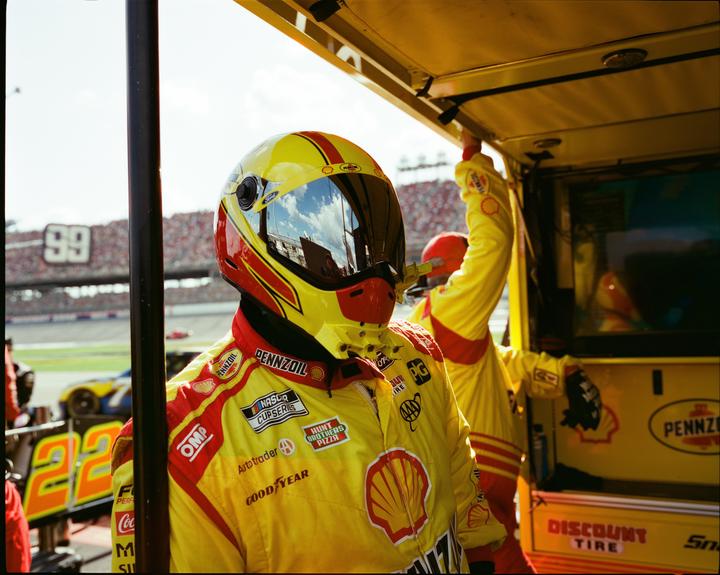 A man in a yellow racing suit and helmet stands in a pit stop area with a race track and spectators in the background.