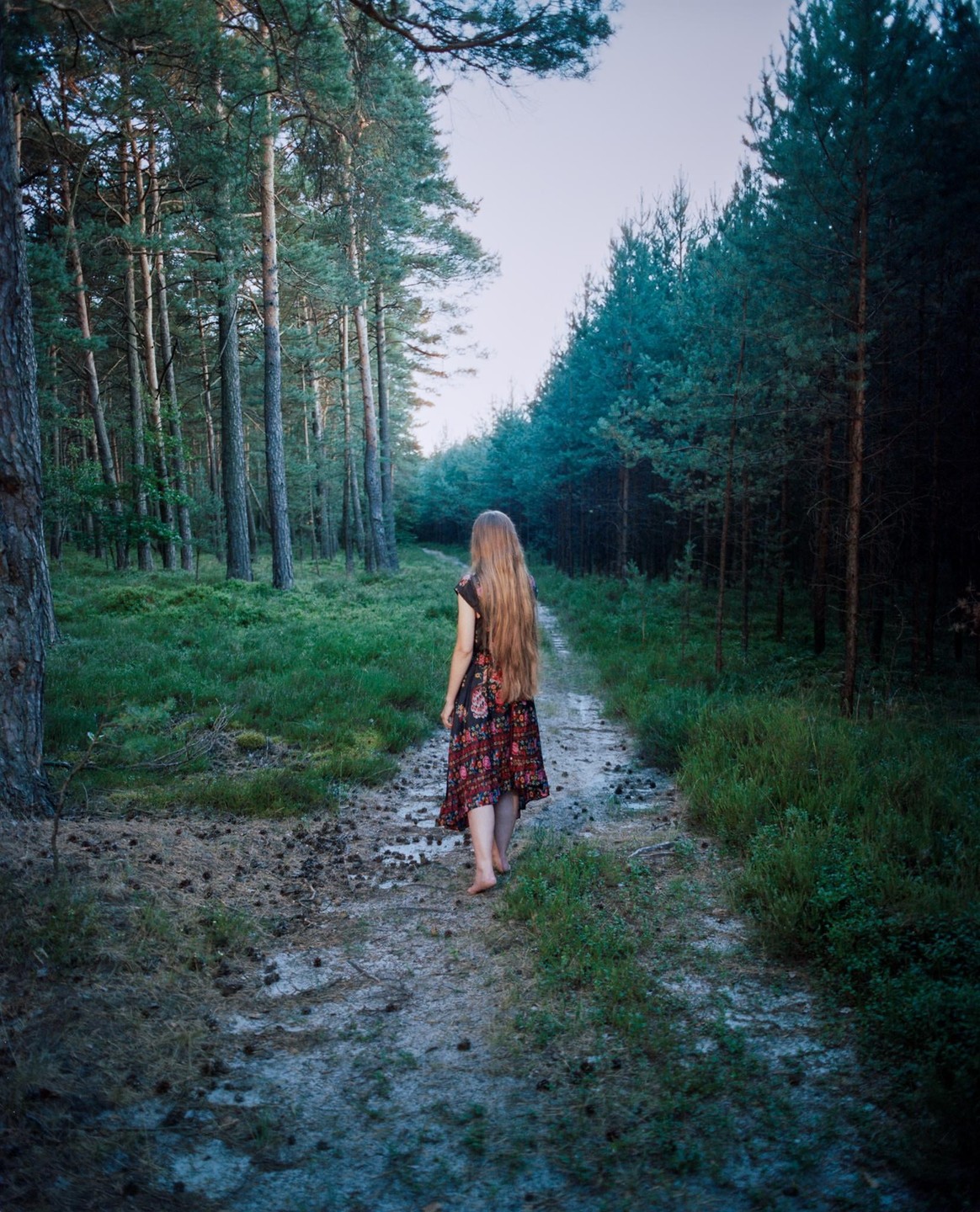 A woman with long hair walks barefoot down a path in a forest.