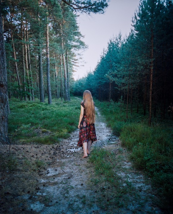 A woman with long hair walks barefoot down a path in a forest.
