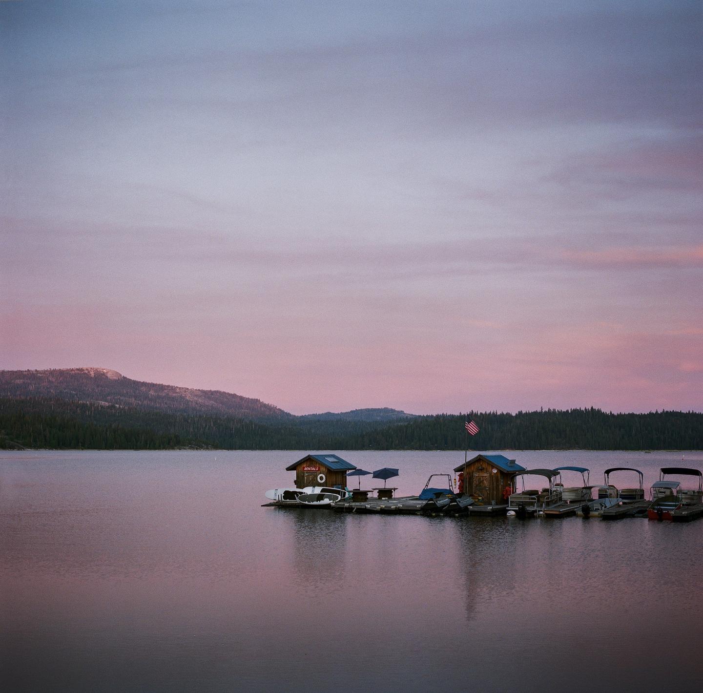 A floating dock with several boats and two small buildings sits on a calm lake surrounded by forested mountains under a pink and purple sky.