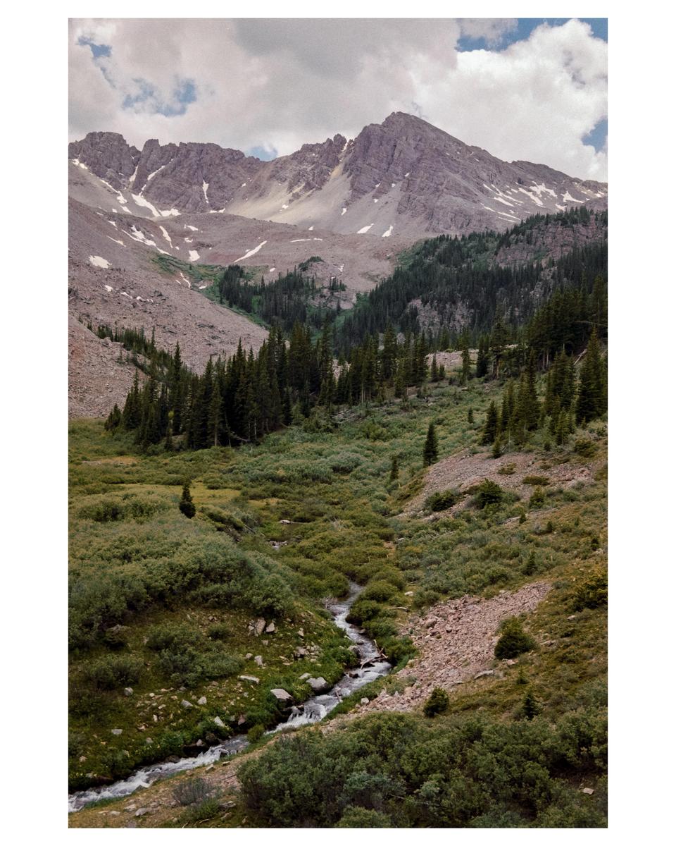 A stream winds through a valley with evergreen trees and mountains under a cloudy sky.