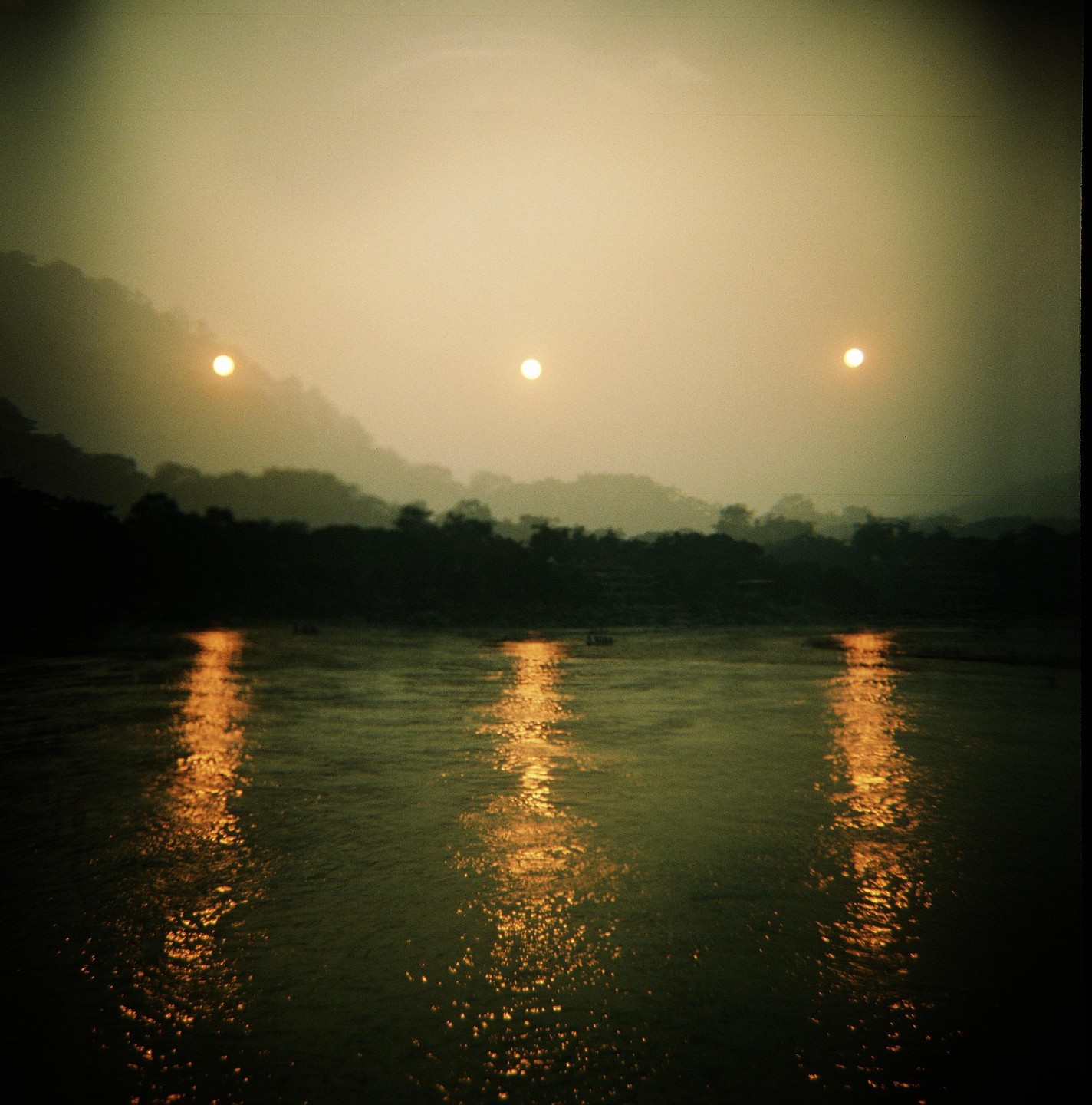Three bright lights are reflected on the surface of a river with a dark, tree-lined bank and hazy hills in the background.