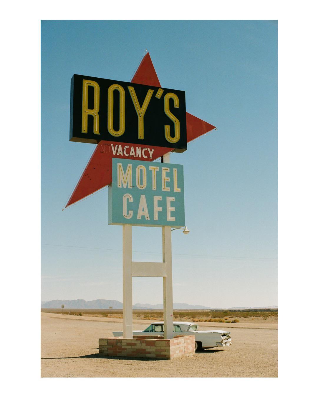 A large sign for Roy's Motel and Cafe stands in a desert landscape with a vintage car parked nearby.