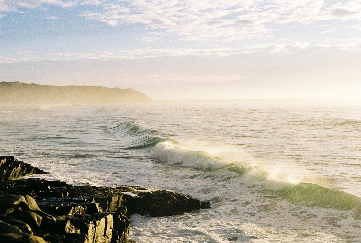 Waves crash against a rocky shore with a hazy headland in the distance under a cloudy sky.