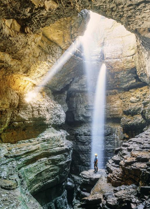 A person stands on a rock formation inside a cave with light beams and a waterfall.