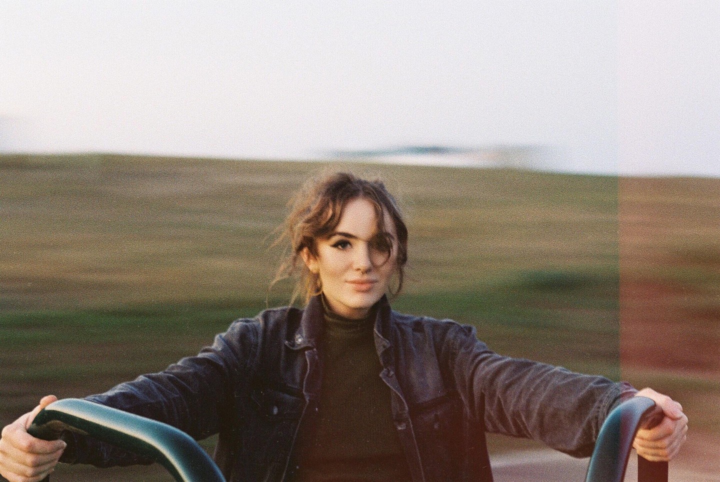 A woman with dark hair and a dark jacket is holding onto a green bar with a blurred field in the background.