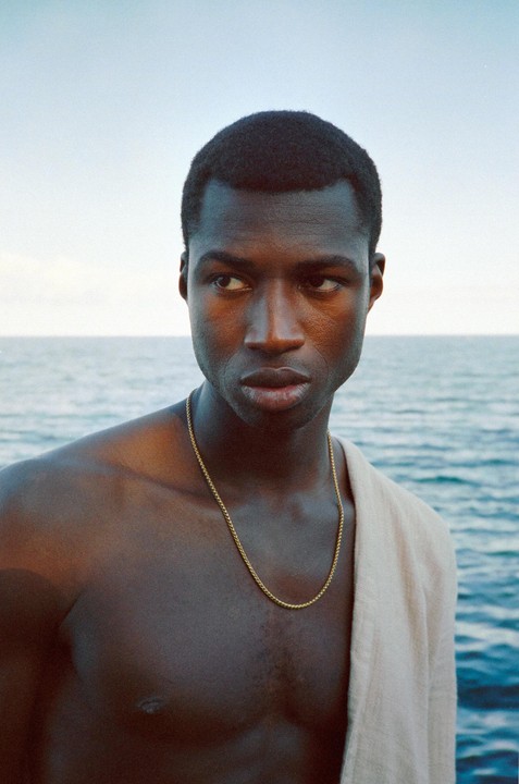 A man with a gold chain around his neck looks to the side with the ocean in the background.