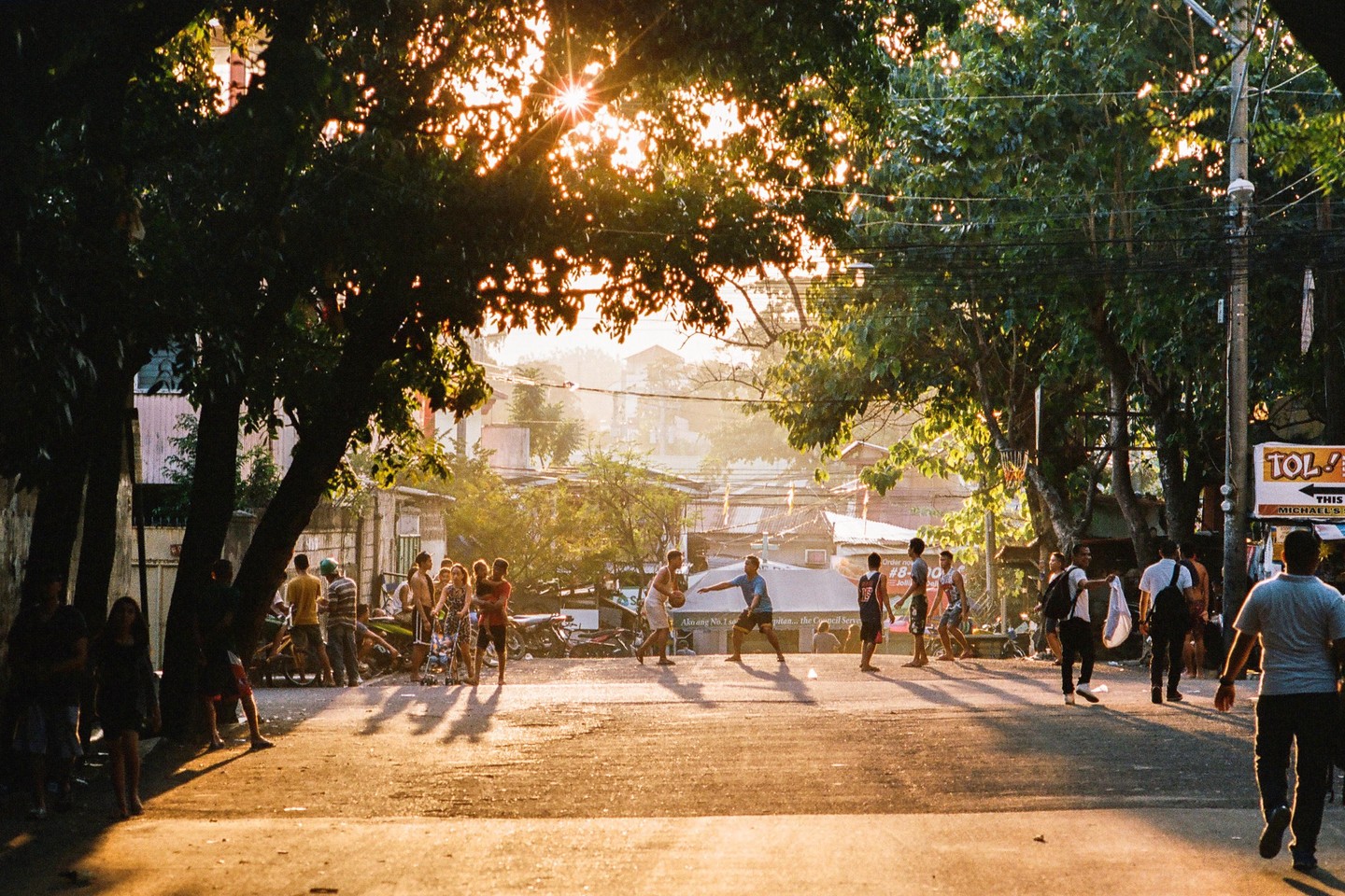 A group of men play basketball on a sunlit street lined with trees and buildings in Manila.