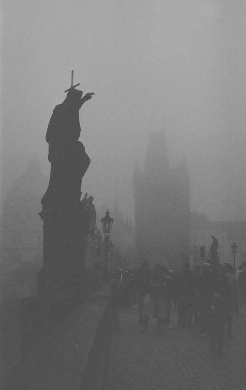 A statue with a cross on its head stands on a bridge with people walking in the fog.