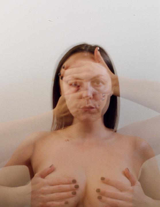 A woman with long dark hair covers her bare chest with her hands while her face is distorted by a double exposure.