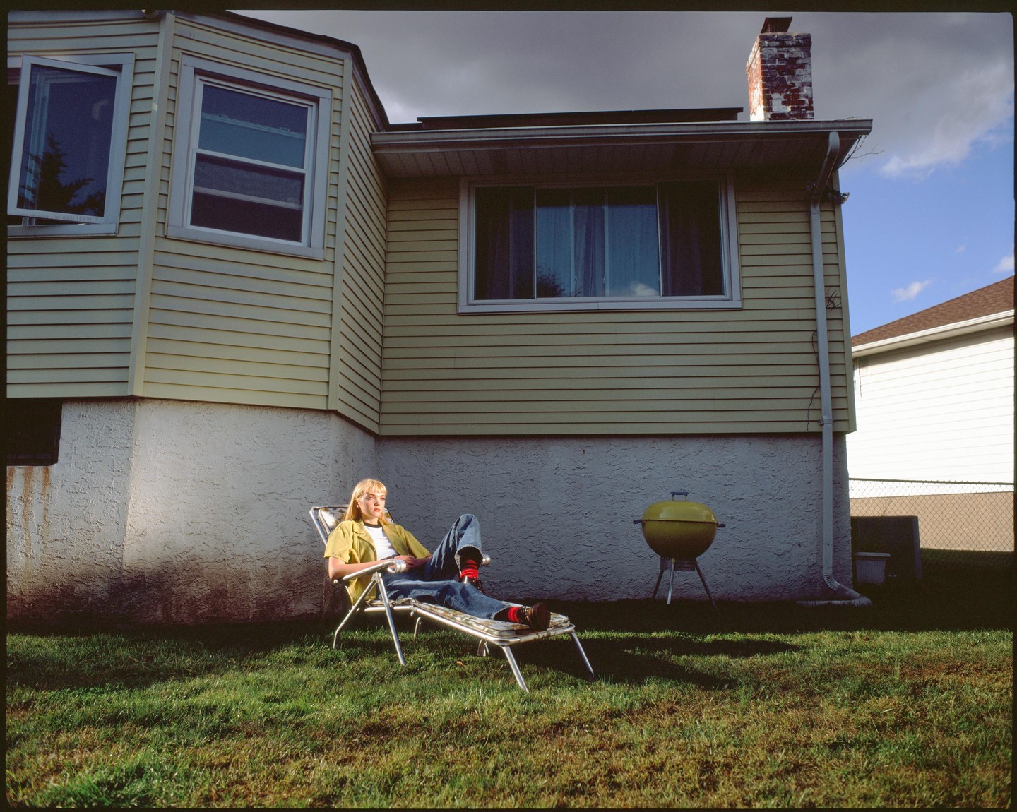A woman sits in a lounge chair in a grassy backyard next to a house with yellow siding and a yellow barbecue.