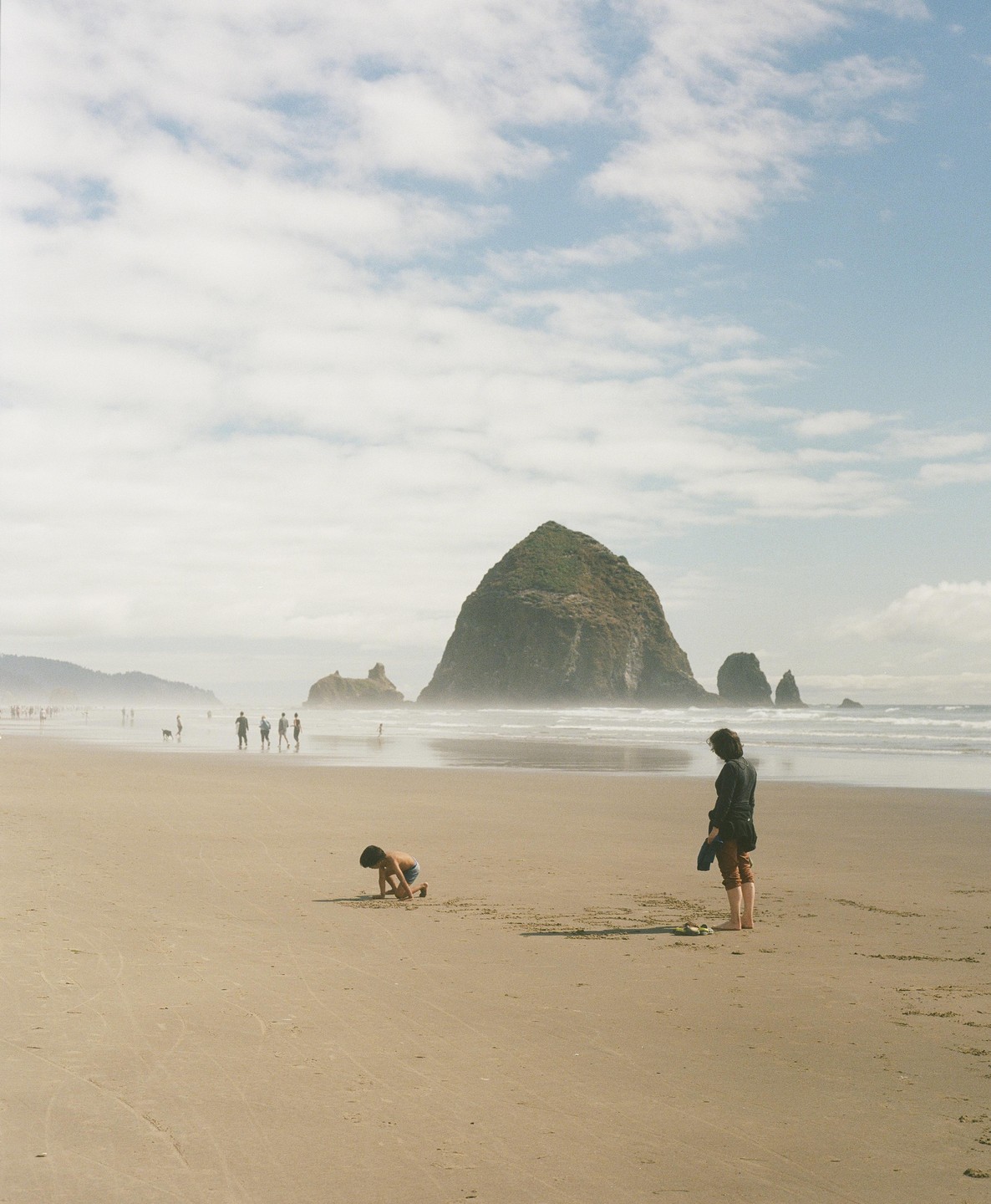 A child plays in the sand on a beach with a large rock formation in the background while a woman stands nearby.