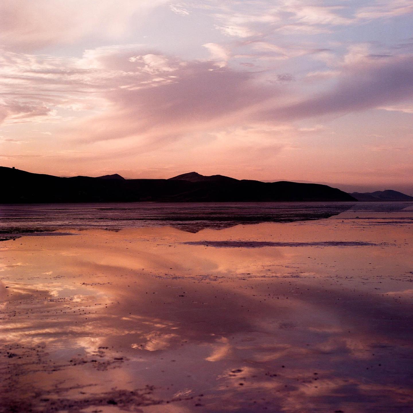 A body of water reflects the pink and purple sky at sunset with mountains in the distance.