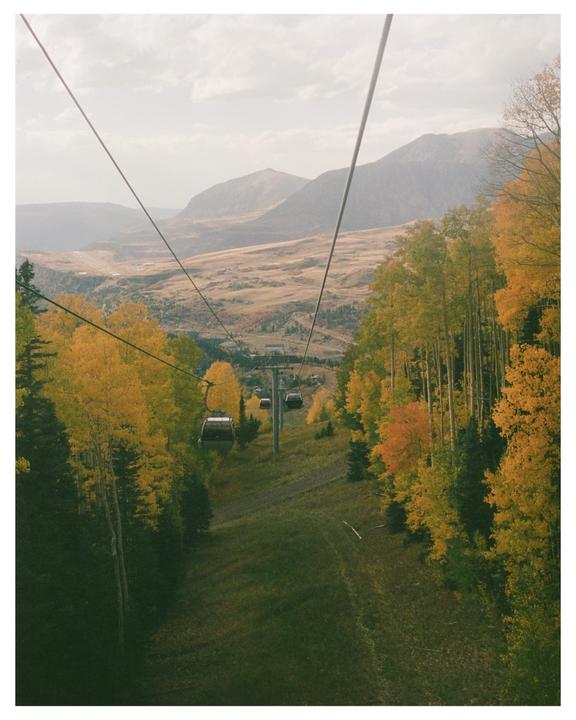 A gondola travels down a mountain slope lined with autumn trees, with a valley and mountains in the background.