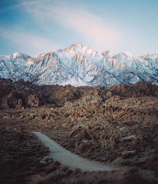 A dirt road leads through a rocky landscape towards snow-capped mountains under a clear sky.