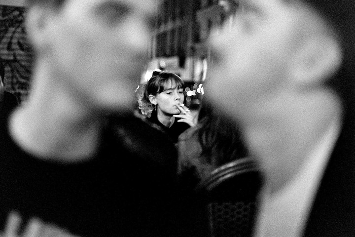A woman with bangs and curly hair smokes a cigarette in a dark setting.