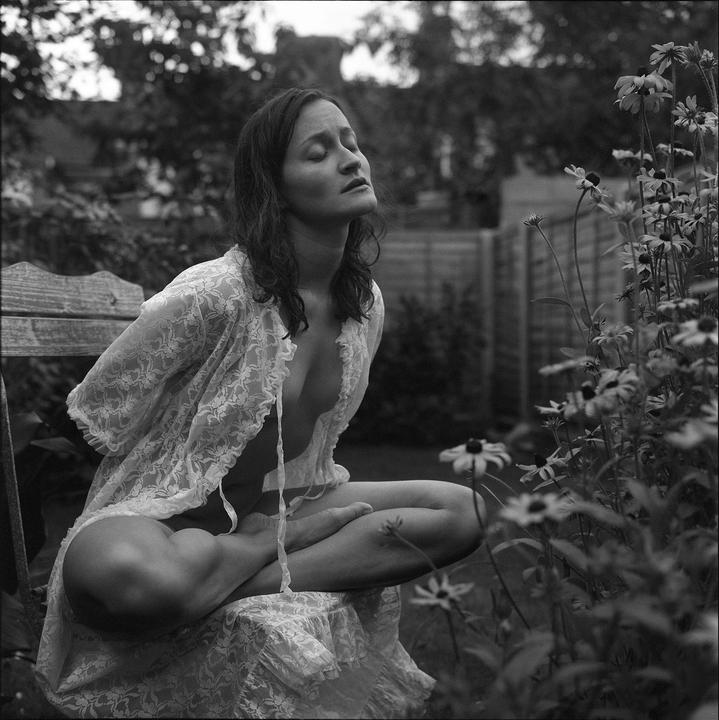 A woman in a lace robe sits on a wooden bench with her eyes closed in a garden.