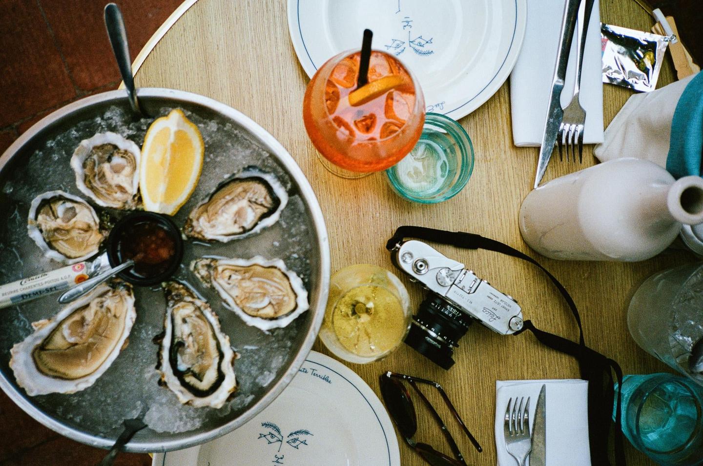 A platter of oysters with lemon and sauce is on a wooden table next to a cocktail, a camera, and other dining items.