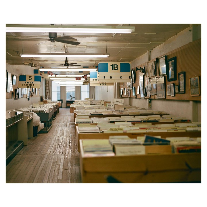 A long room with wooden floors and fluorescent lights is filled with rows of records in wooden bins.