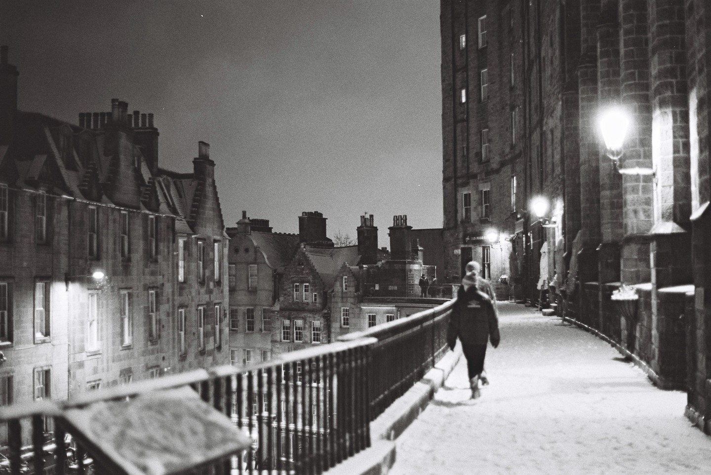 A person walks along a snow-covered street lined with buildings at night.