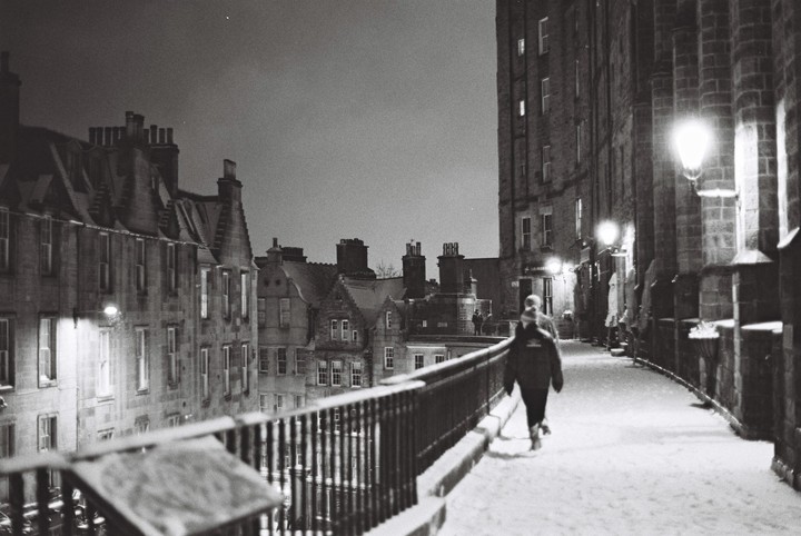 A person walks along a snow-covered street lined with buildings at night.